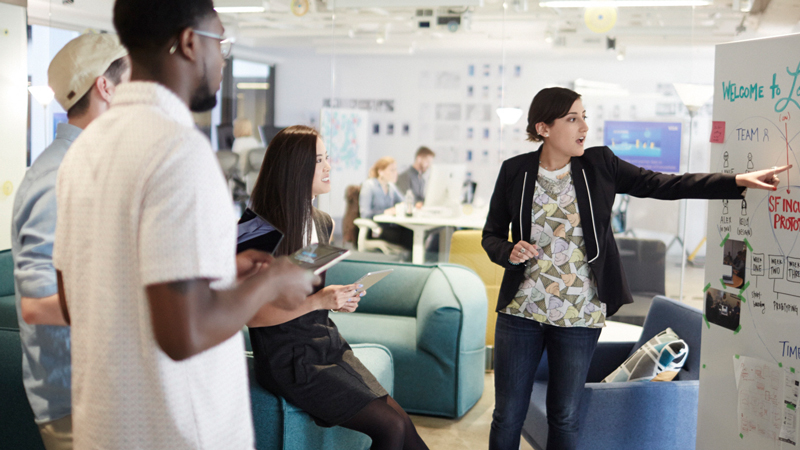 Three people looking at a woman pointing at a diagram on a whiteboard.
