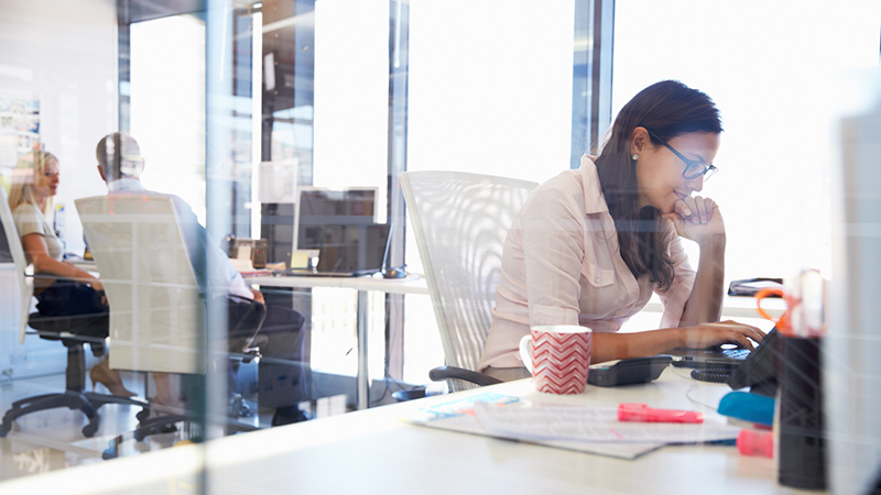 Marquee-VE-OfficeWoman_800x450 Woman working at her desk in an office with people in the background chatting at their desks.