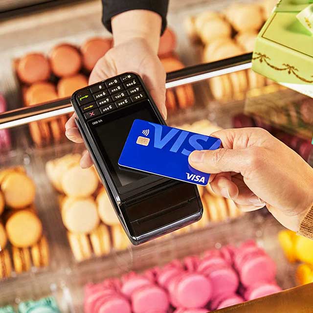 A display of cookies is visible in the background as a person holds up a Visa contactless payment card for payment. 